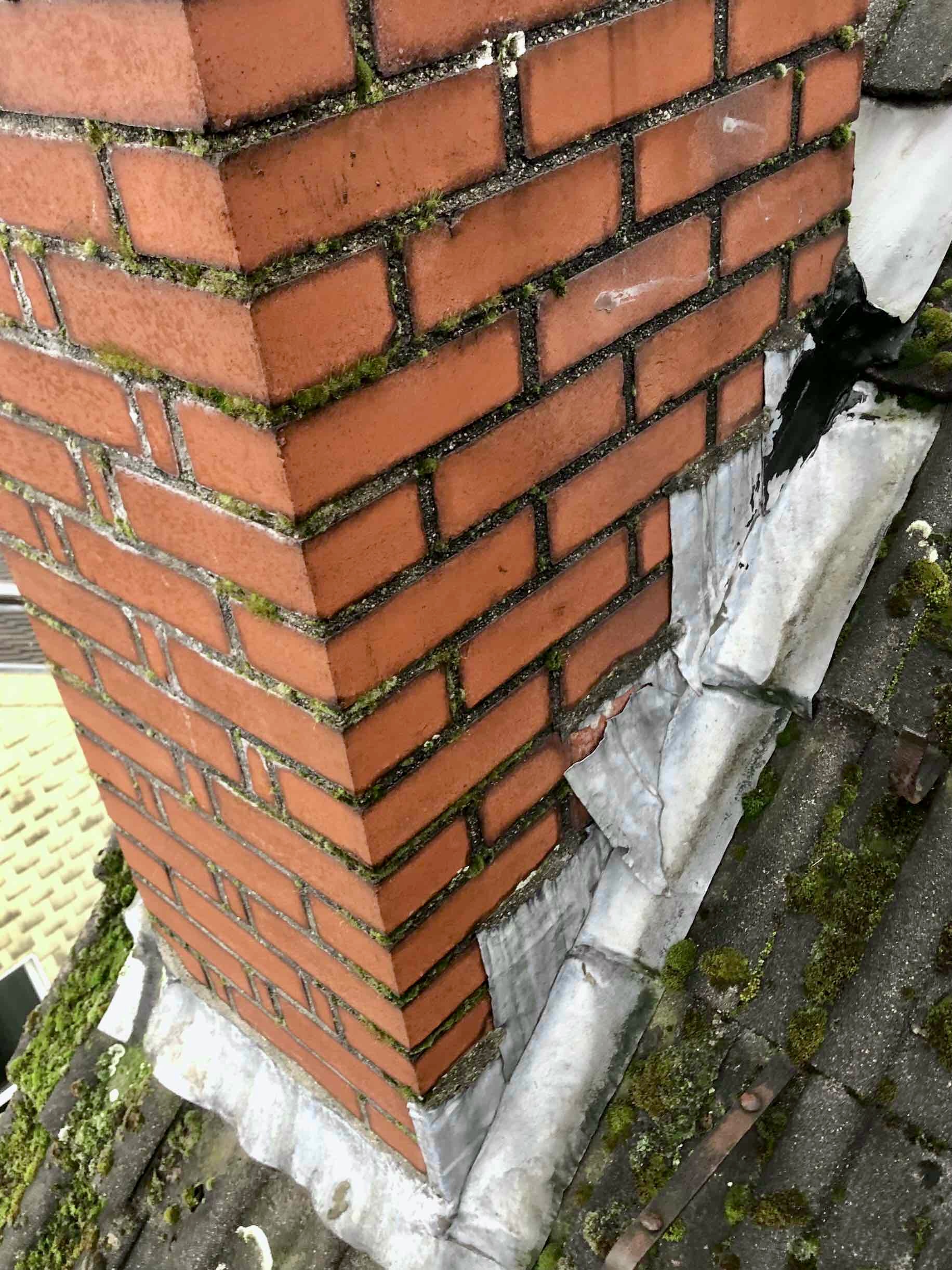 A red brick chimney with moss in its mortar joints and flashing lifting at one corner